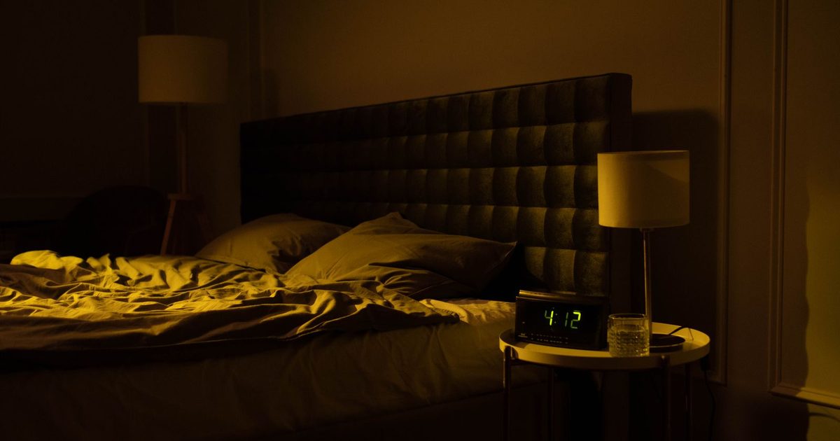A dimly lit bedroom with a bedside lamp glowing softly in the evening