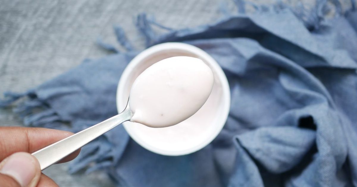 A hand holding a spoonful of plain yoghurt above a bowl on a blue cloth